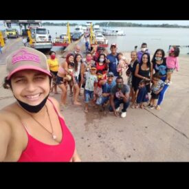 Mulher parda sorrindo em primeiro plano, ela usa blusa regata na cor rosa e boné rosa, ao fundo um grupo de mulheres e homens com crianças de colo e crianças pequenas, alguns estão de máscara descartável no queixo, atrás do grupo uma balsa