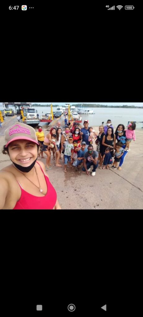 Mulher parda sorrindo em primeiro plano, ela usa blusa regata na cor rosa e boné rosa, ao fundo um grupo de mulheres e homens com crianças de colo e crianças pequenas, alguns estão de máscara descartável no queixo, atrás do grupo uma balsa