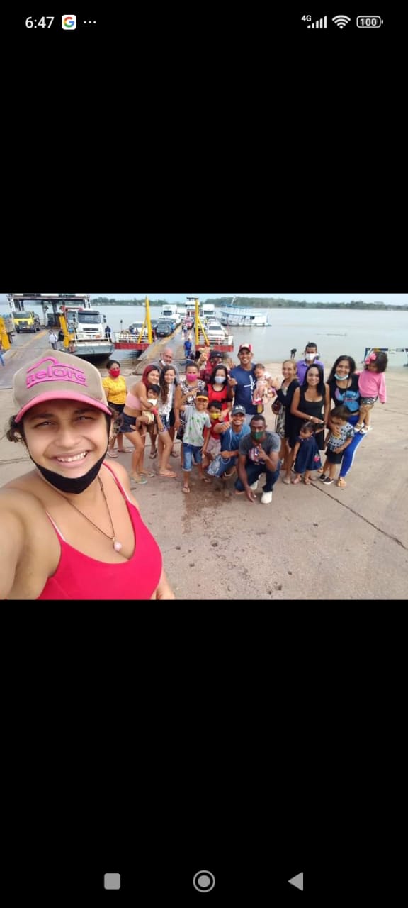 Mulher parda sorrindo em primeiro plano, ela usa blusa regata na cor rosa e boné rosa, ao fundo um grupo de mulheres e homens com crianças de colo e crianças pequenas, alguns estão de máscara descartável no queixo, atrás do grupo uma balsa